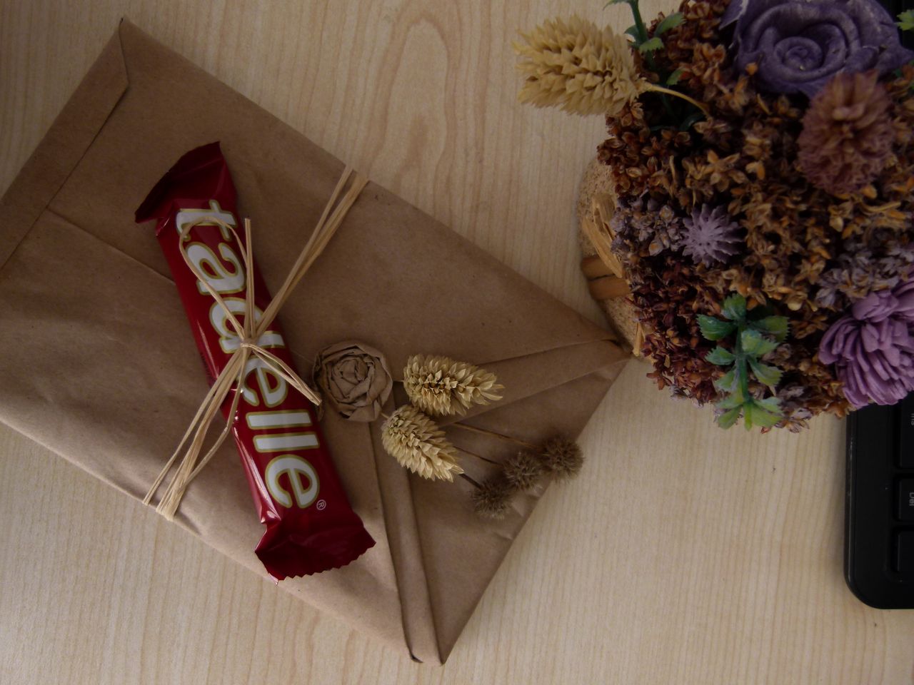 A rustic gift wrapped with dried flowers, brown paper, and a chocolate bar on a wooden table.
